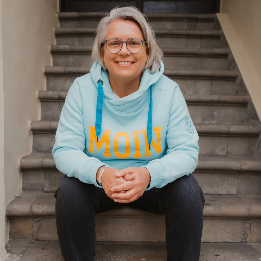 Kirsten Biema sitzt lächelnd auf einer Steintreppe, im hellblauen MOIN-Hoodie